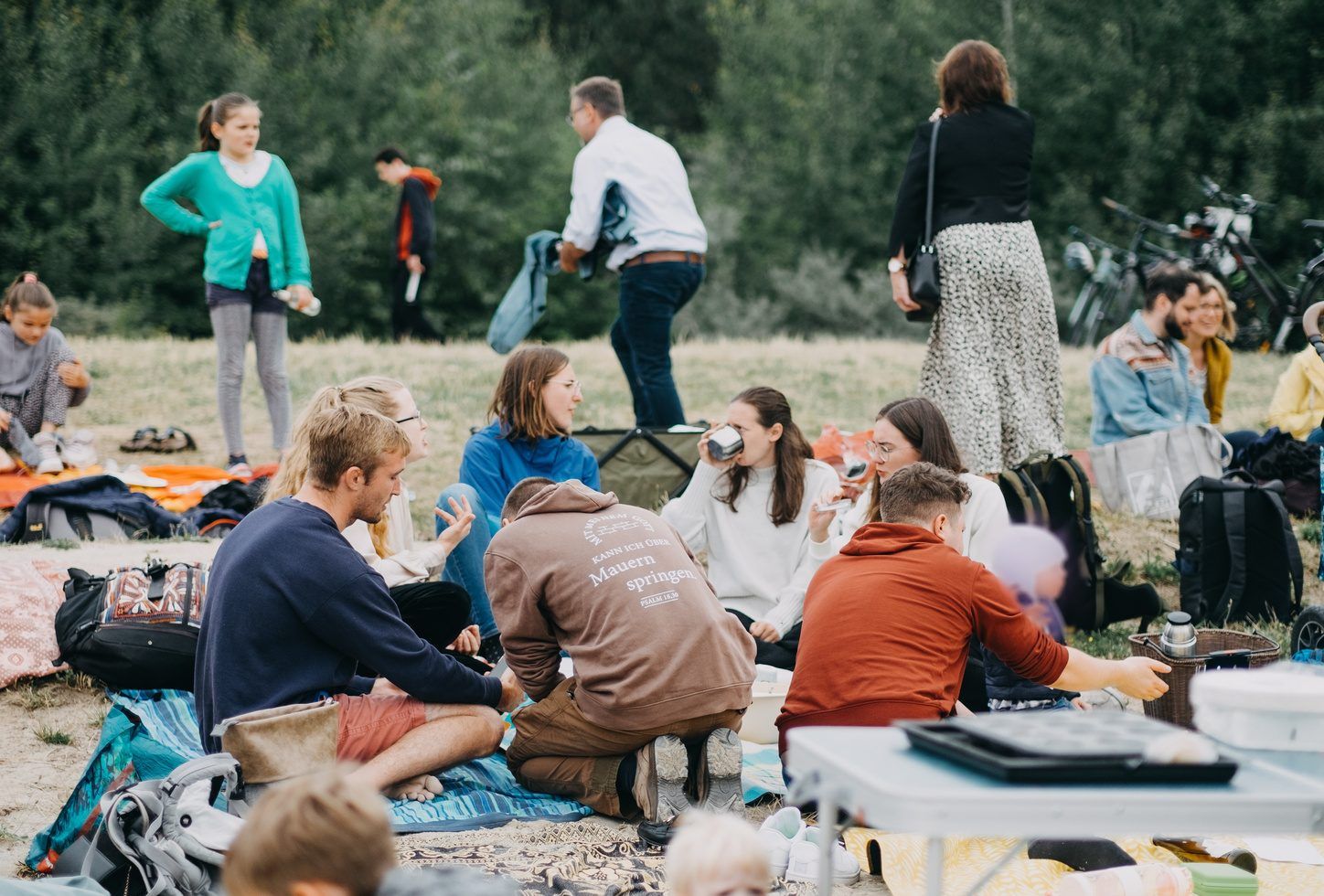 Eine Gruppe junger Menschen sitzt auf Decken auf einer Wiese und unterhält sich, im Hintergrund Bäume und Fahrräder