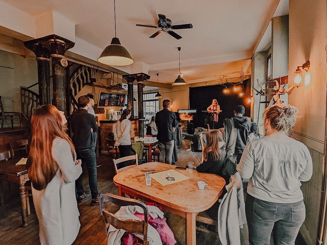 Menschen stehen in einem Café mit Holztischen und Hängelampen und hören einer Sängerin mit Gitarre auf einer kleinen Bühne zu