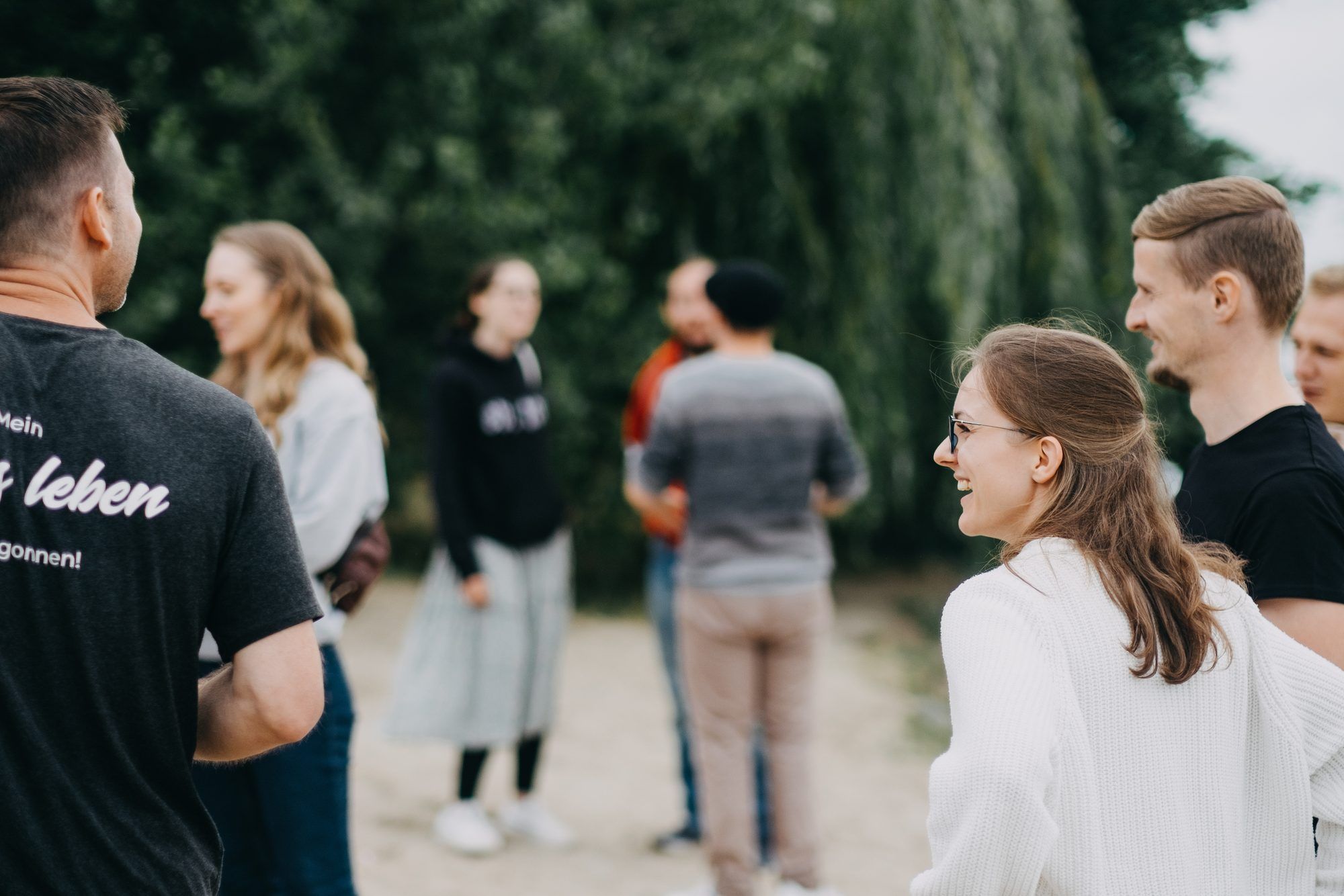 Eine junge Frau mit Brille lacht im Gespräch mit einer Gruppe junger Erwachsener, die vor Bäumen im Freien steht