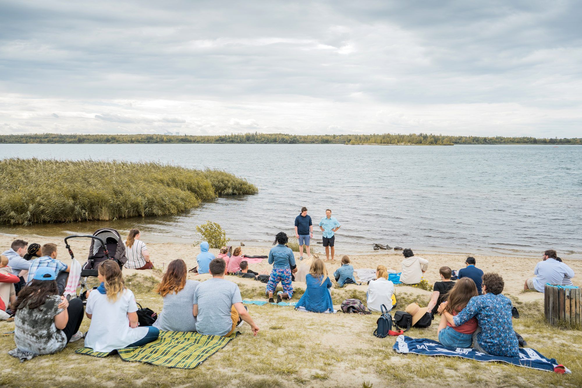 Eine Gruppe sitzt auf Decken am Sandstrand eines Sees und hört zwei Männern zu, die am Ufer stehen