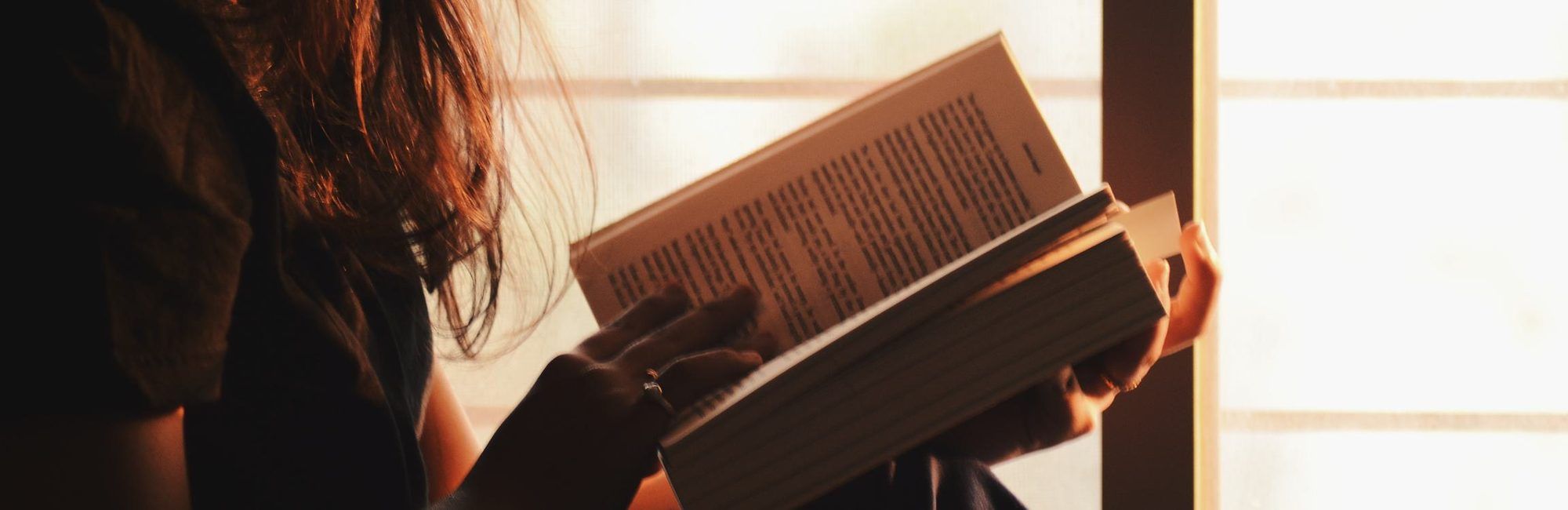 Eine Frau sitzt an einem hellen Fenster und liest in einem Buch