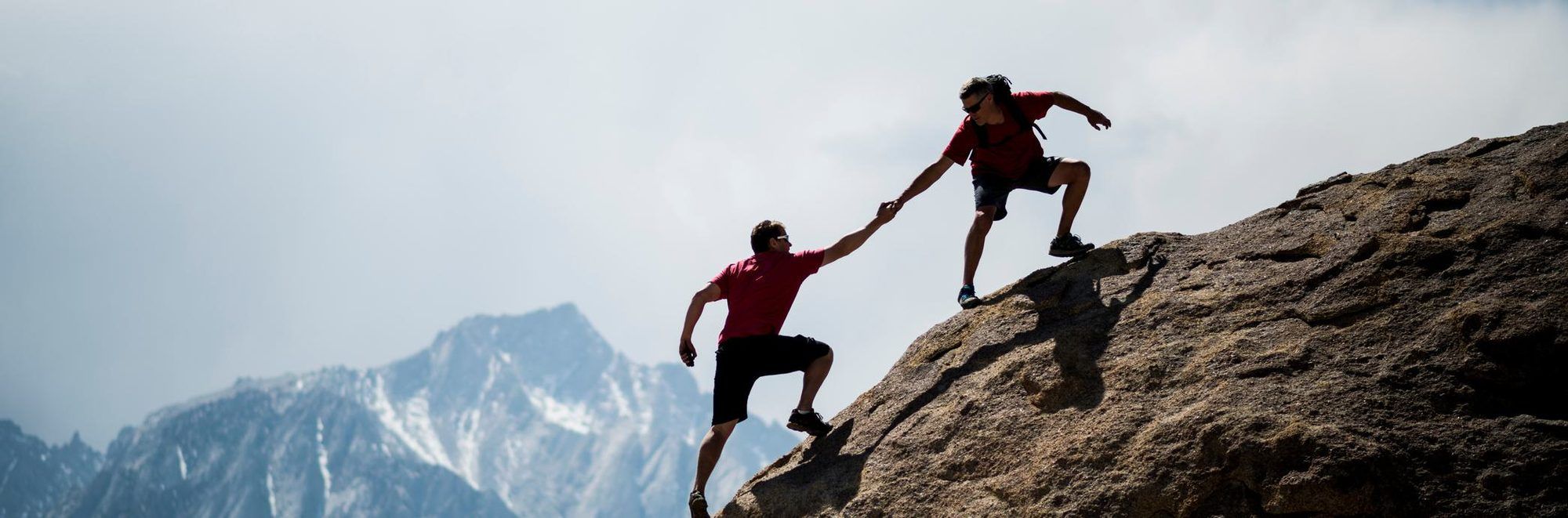 Ein Bergsteiger reicht einem anderen an einem steilen Felsen die Hand und zieht ihn hoch, im Hintergrund schneebedeckte Berge