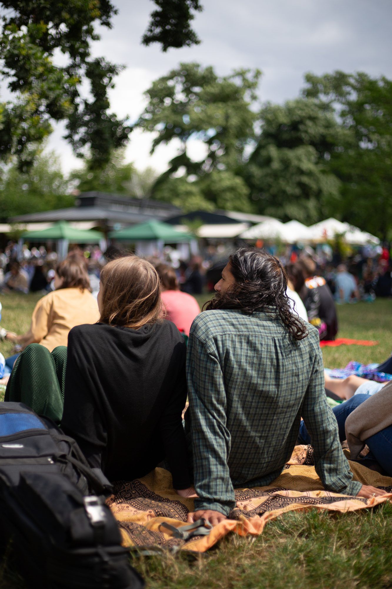 Zwei Menschen sitzen von hinten gesehen auf einer Decke im Gras, vor ihnen viele weitere Menschen und Zelte in einem Park