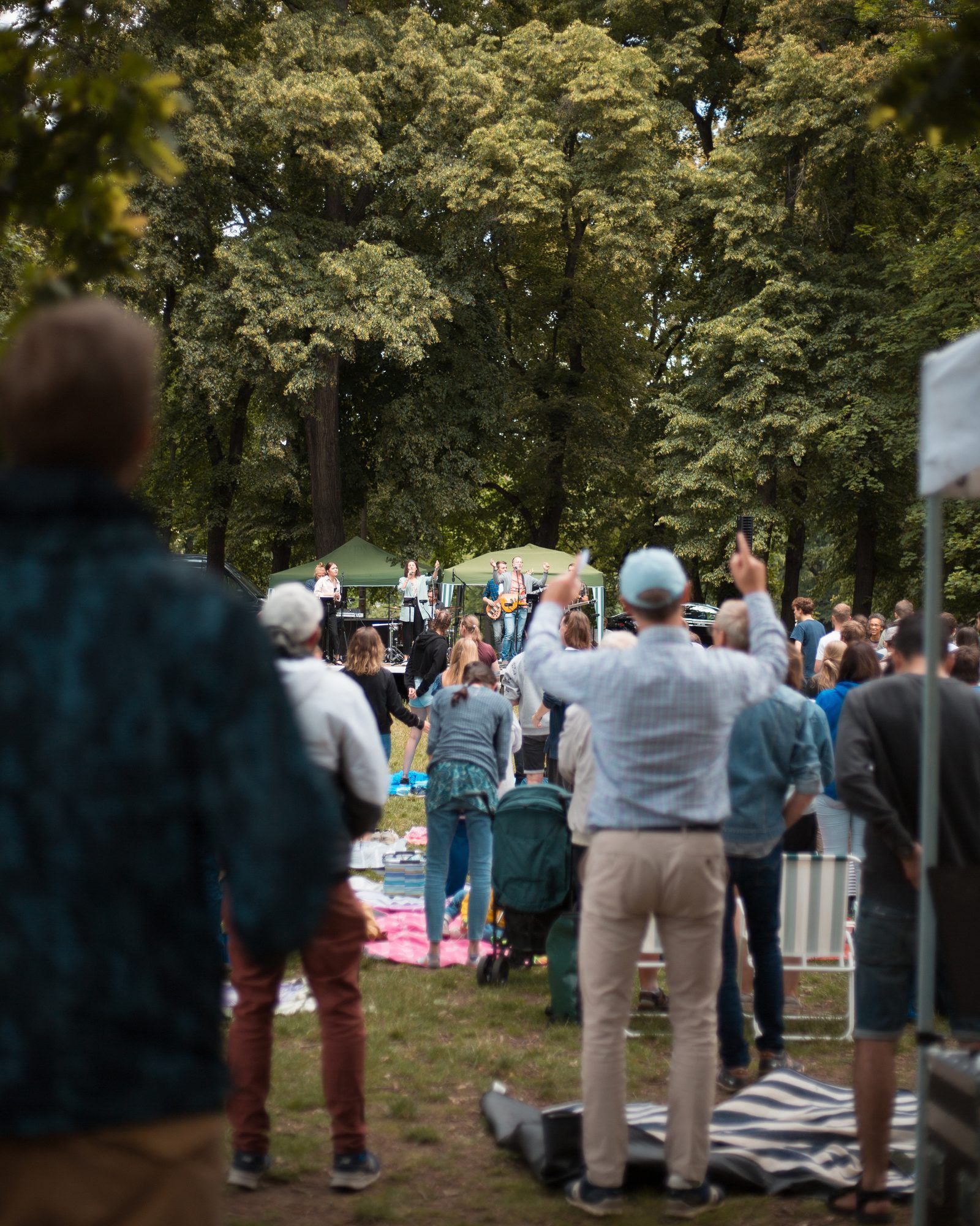 Menschen feiern im Park einen Open-Air-Gottesdienst, ein Mann vorne hebt die Hände, im Hintergrund spielt eine Band unter grünen Pavillons