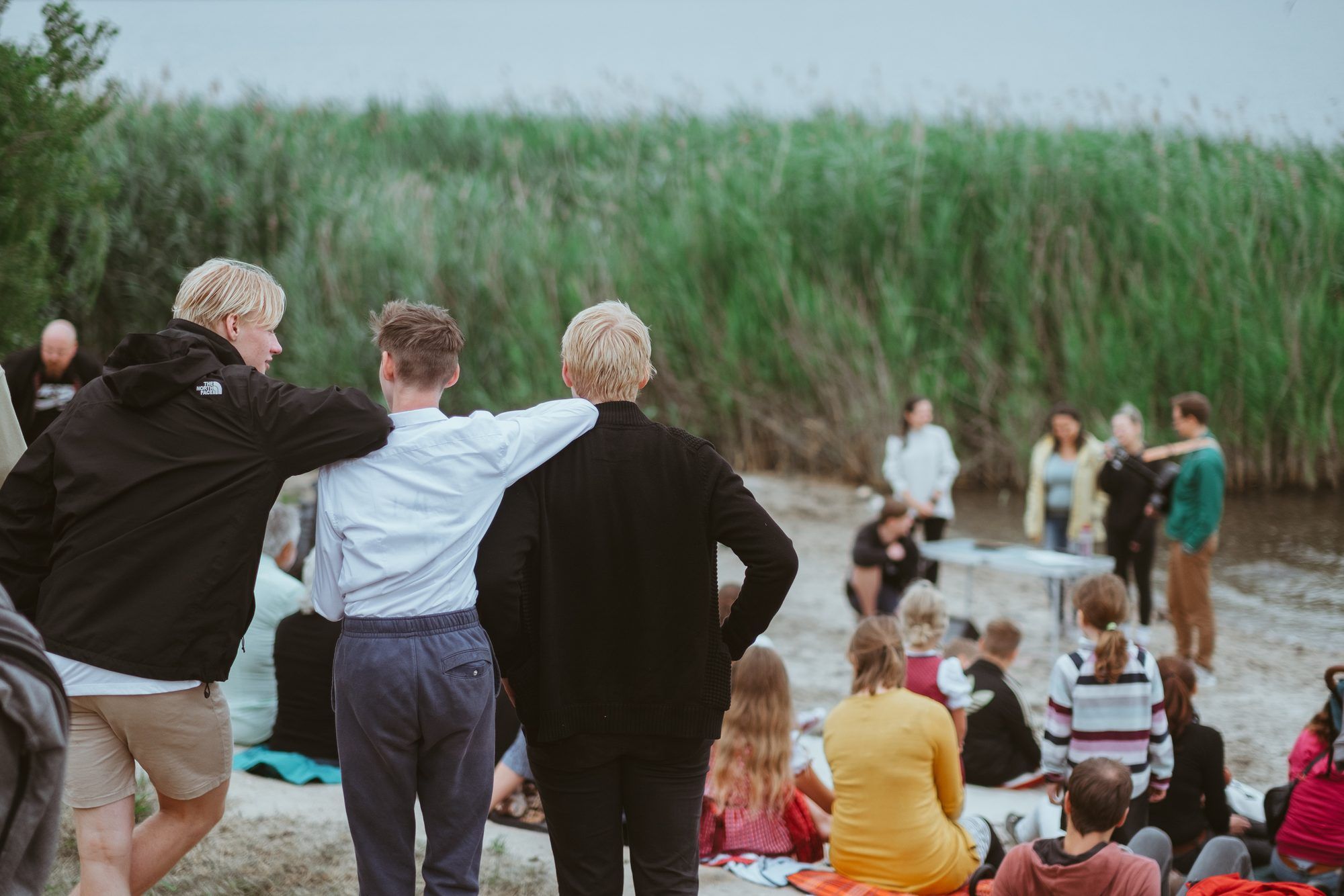 Drei junge Männer stehen Arm in Arm am Ufer vor hohem Schilf und schauen einer Gruppe zu, die auf dem Sand sitzt