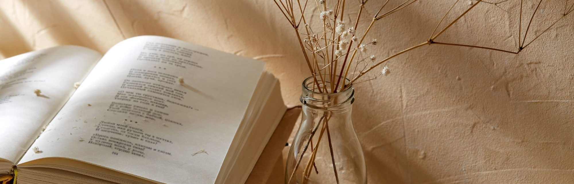 Aufgeschlagenes Gedichtbuch neben einer Glasflasche mit getrockneten Blumen vor einer beigen Wand