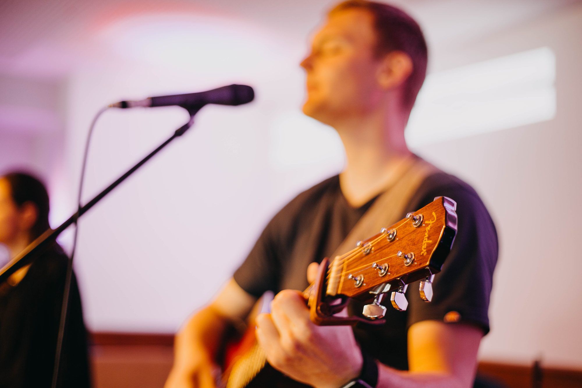 Ein Mann spielt Akustikgitarre und singt vor einem Mikrofon, im Vordergrund scharf der Gitarrenkopf