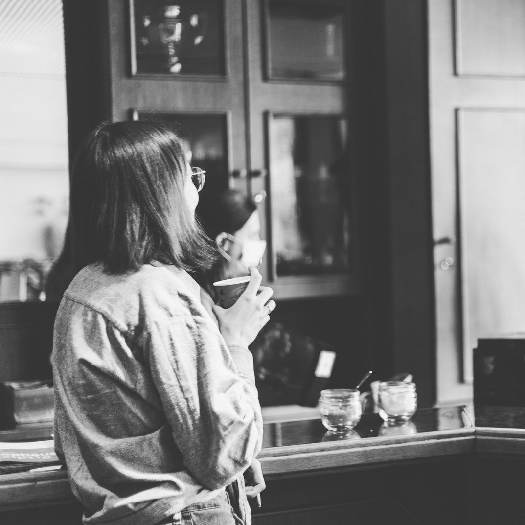 Schwarz-Weiß-Foto einer Frau mit Brille, die an einer Theke steht und aus einer Tasse trinkt
