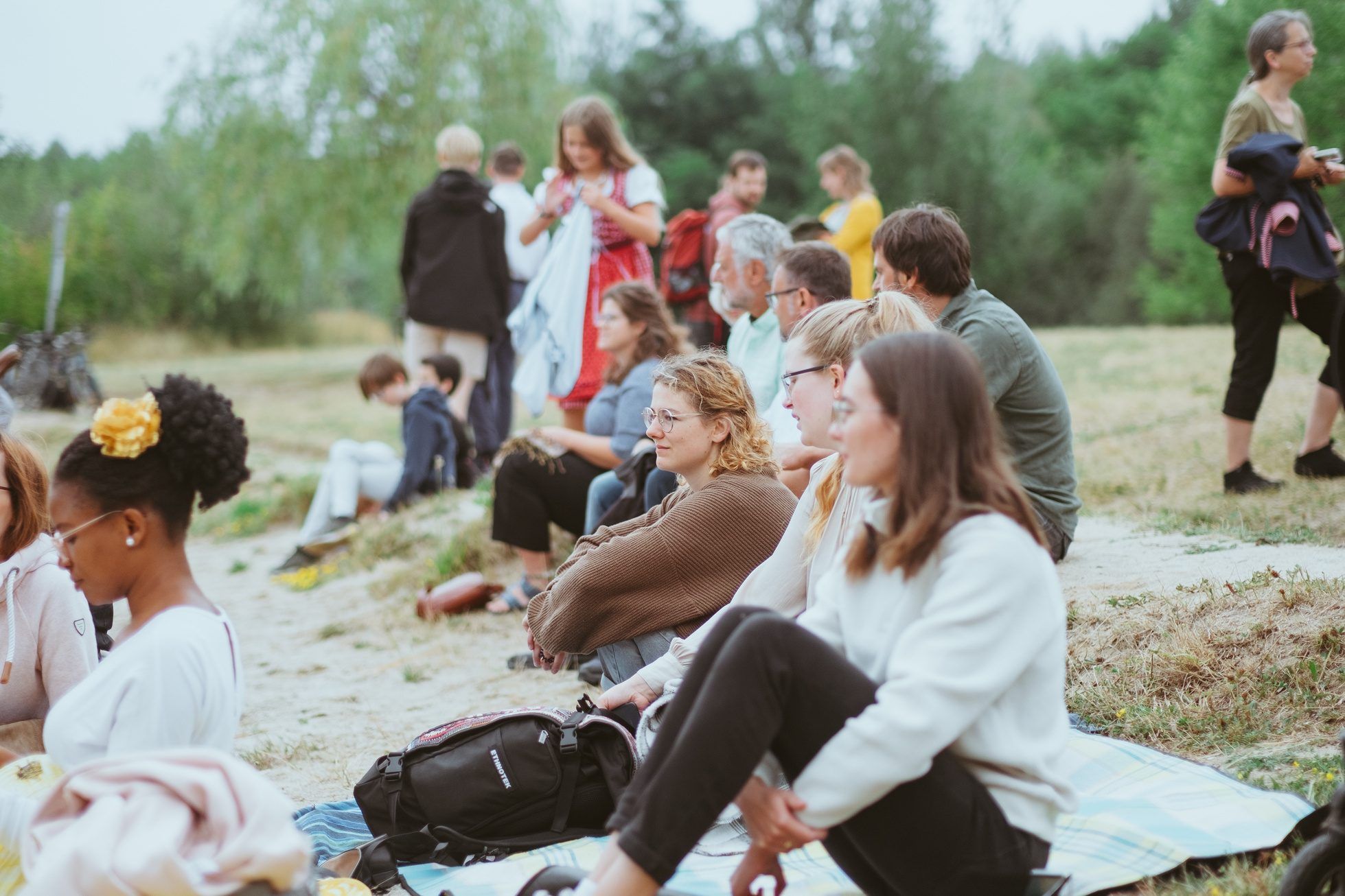 Menschen sitzen auf Decken an einem sandigen Hang im Freien und hören zu, dahinter stehen weitere Personen vor Bäumen