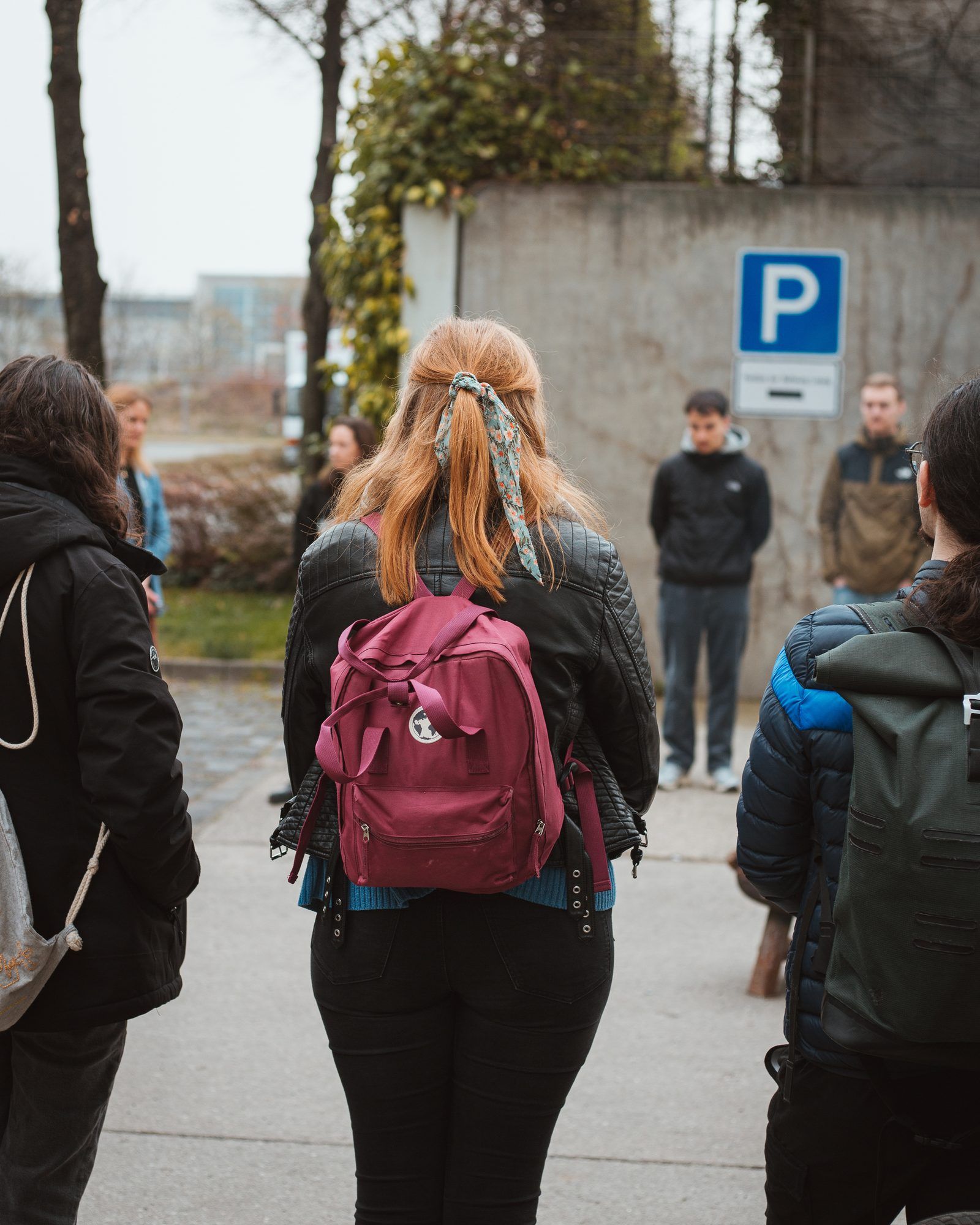 Eine junge Frau mit rotblondem Haar und rotem Rucksack steht mit anderen Menschen im Kreis vor einer Betonmauer