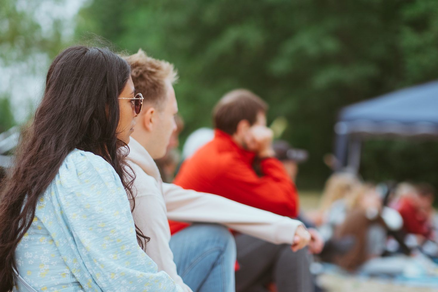 Eine Frau mit Sonnenbrille und ein Mann sitzen im Freien im Gras, im Hintergrund weitere Menschen und Bäume