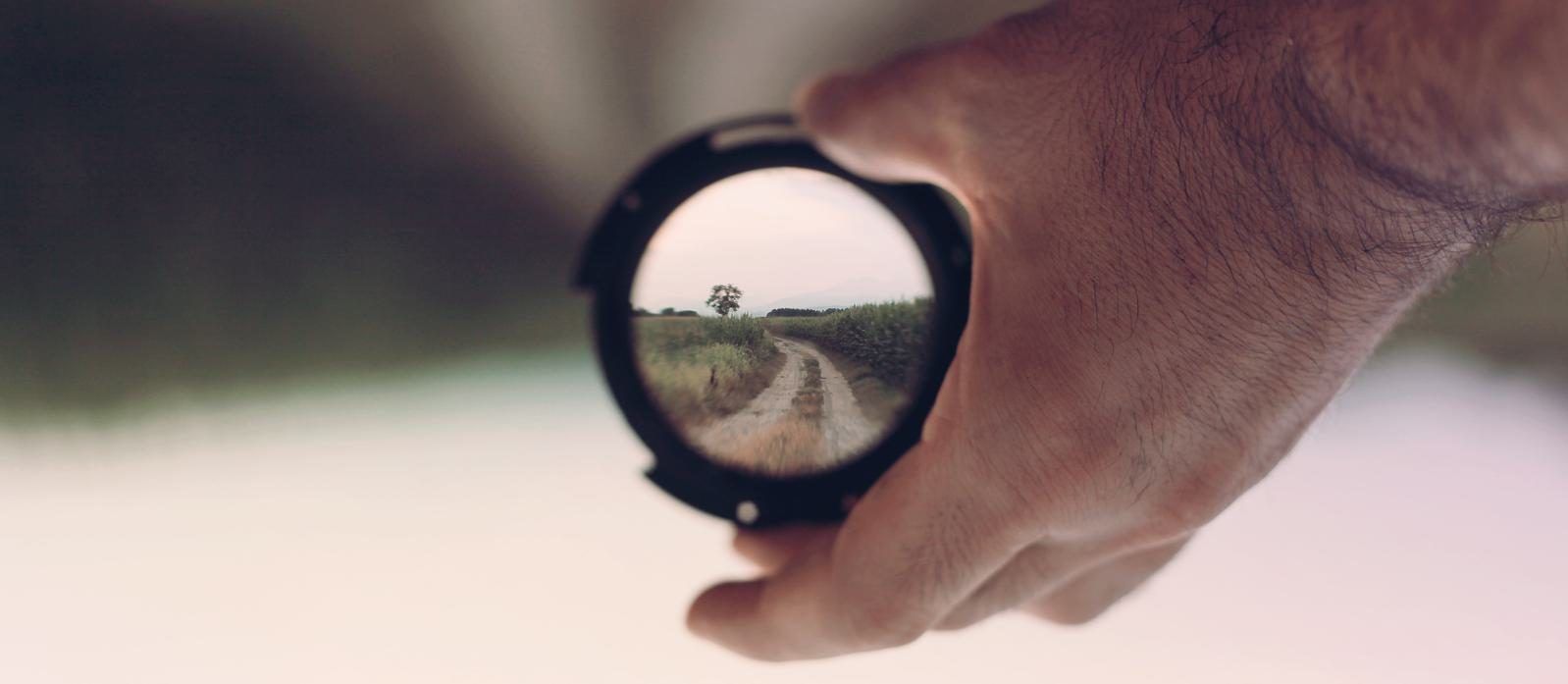 Eine Hand hält ein Objektiv, durch das scharf ein Feldweg mit einem einzelnen Baum zu sehen ist, die Umgebung ist unscharf