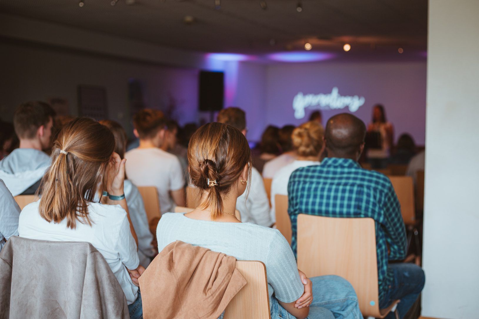 Menschen sitzen von hinten gesehen auf Holzstühlen in einem Gottesdienstraum, vorne eine Sprecherin vor einer violett beleuchteten Wand