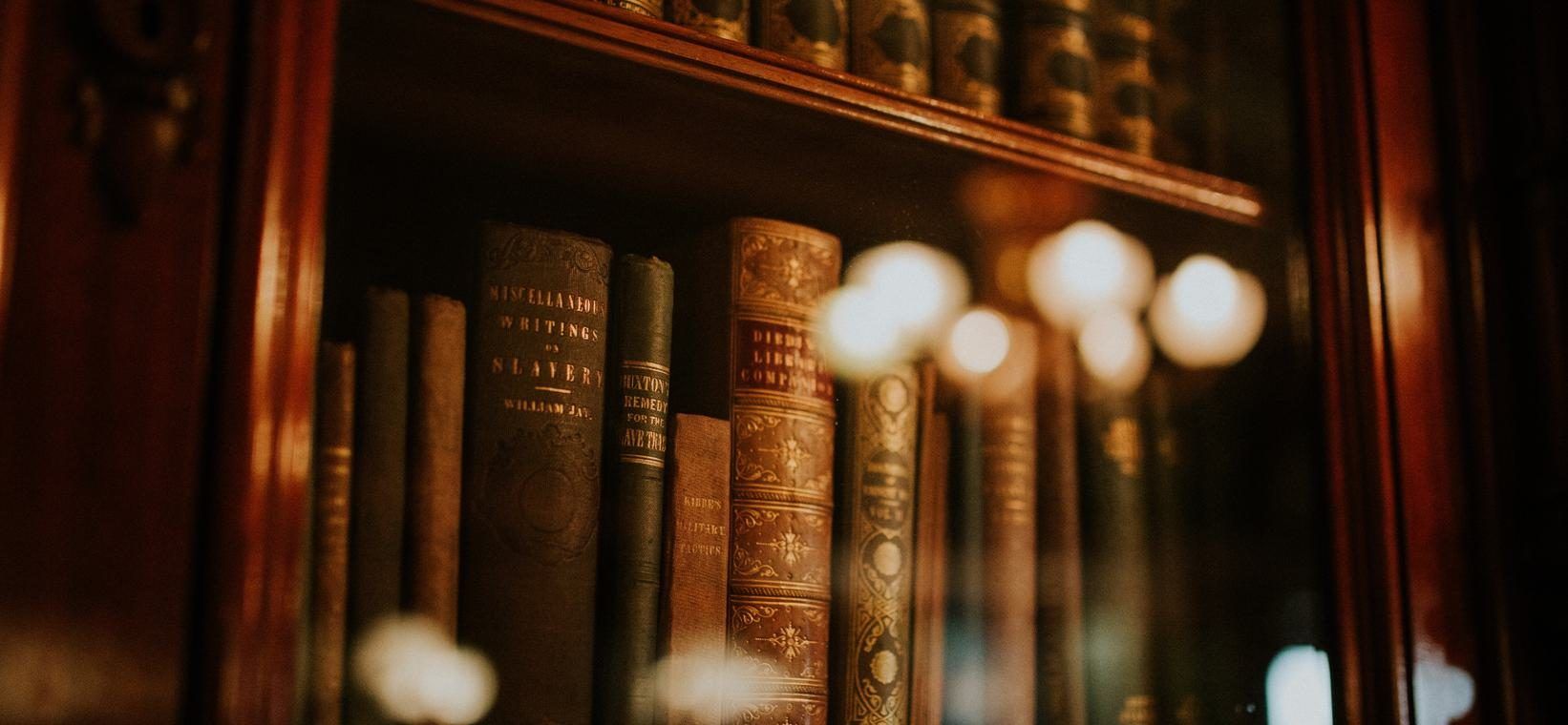 Alte, in Leder gebundene Bücher in einem Holzregal hinter einer Glasscheibe, davor unscharfe Lichtpunkte