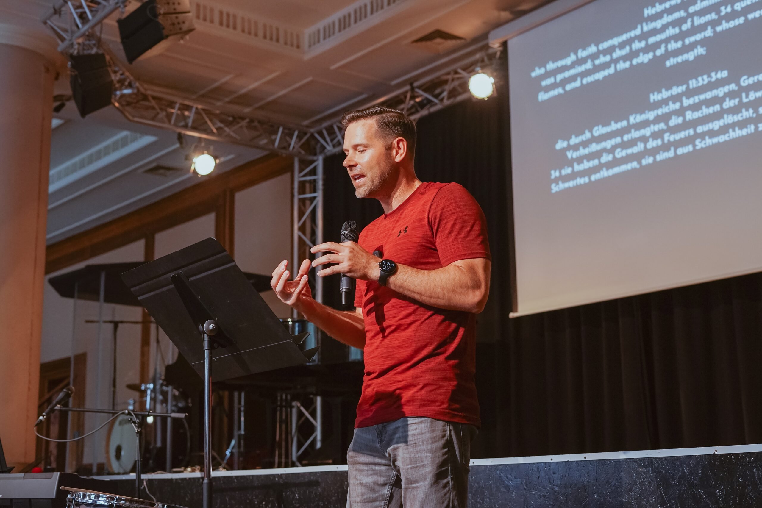 A pastor in a red T-shirt preaching on stage, with a Bible verse on the screen behind him