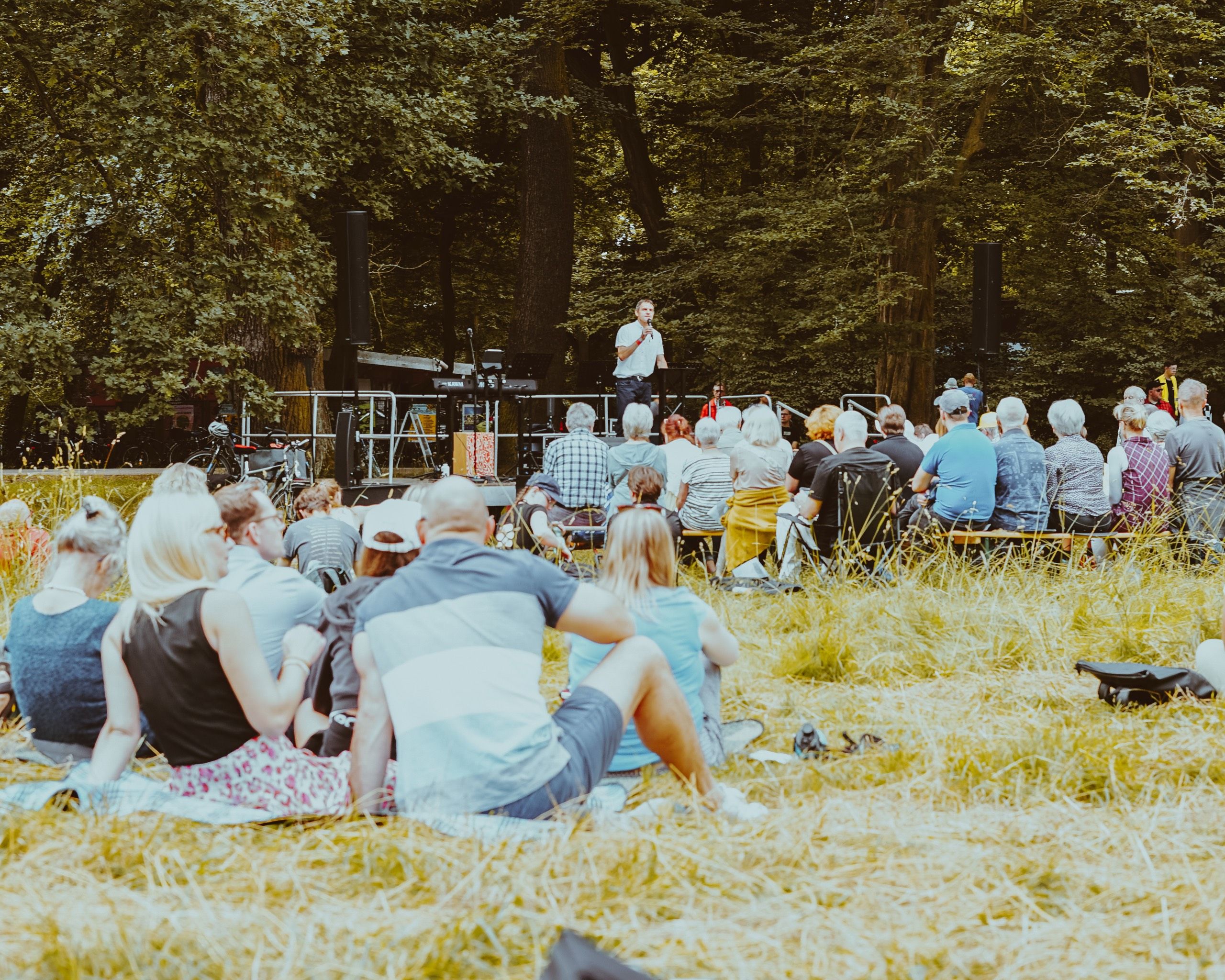 People sitting together on the grass in a park, listening to a speaker at an open-air event